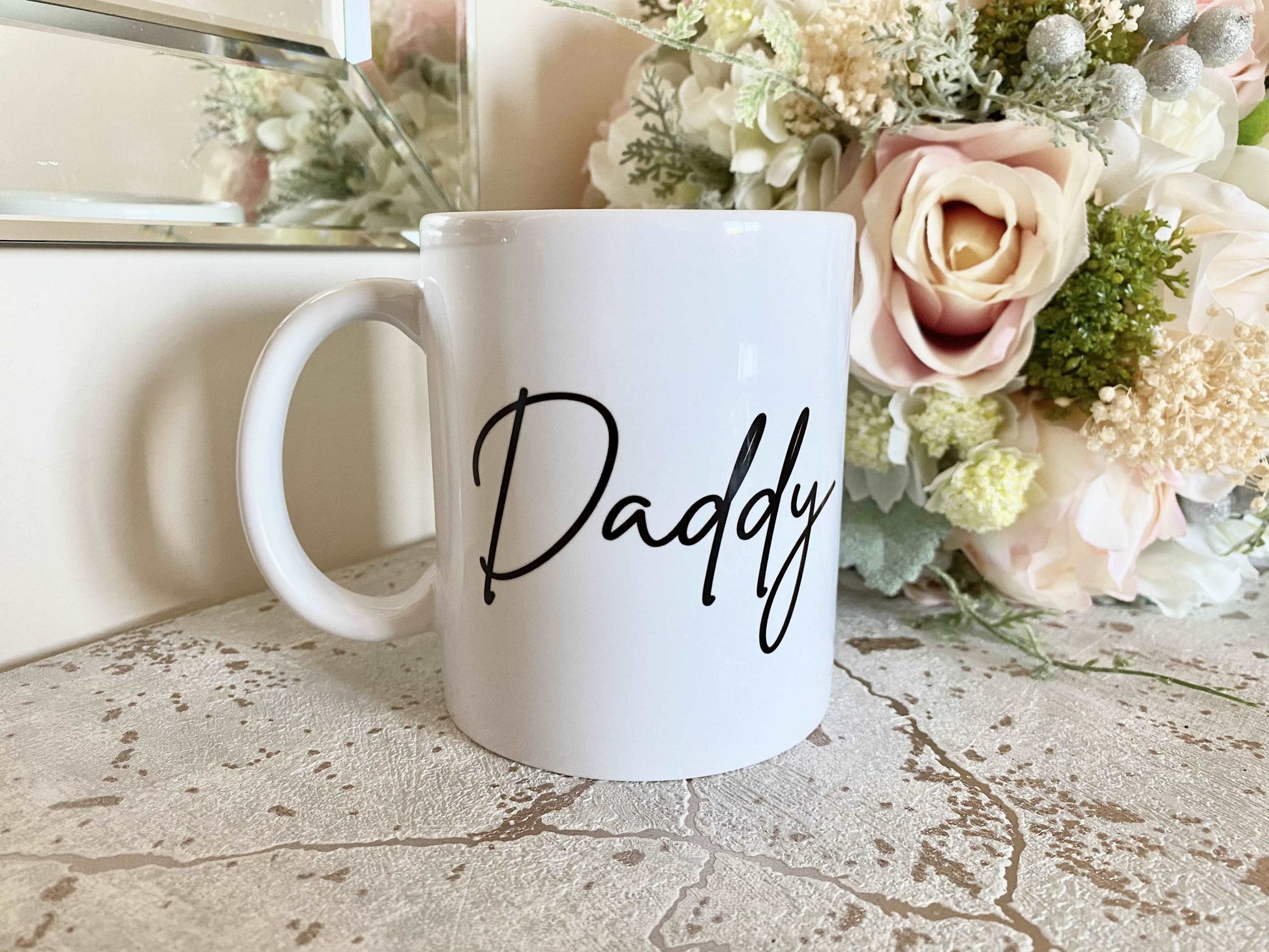 white mug with daddy printed on in a script font. the mug is on a marble effect surface with a flower bouquet behind