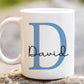 white mug sitting on a stone with a large blue letter D and the name David printed over in a script font