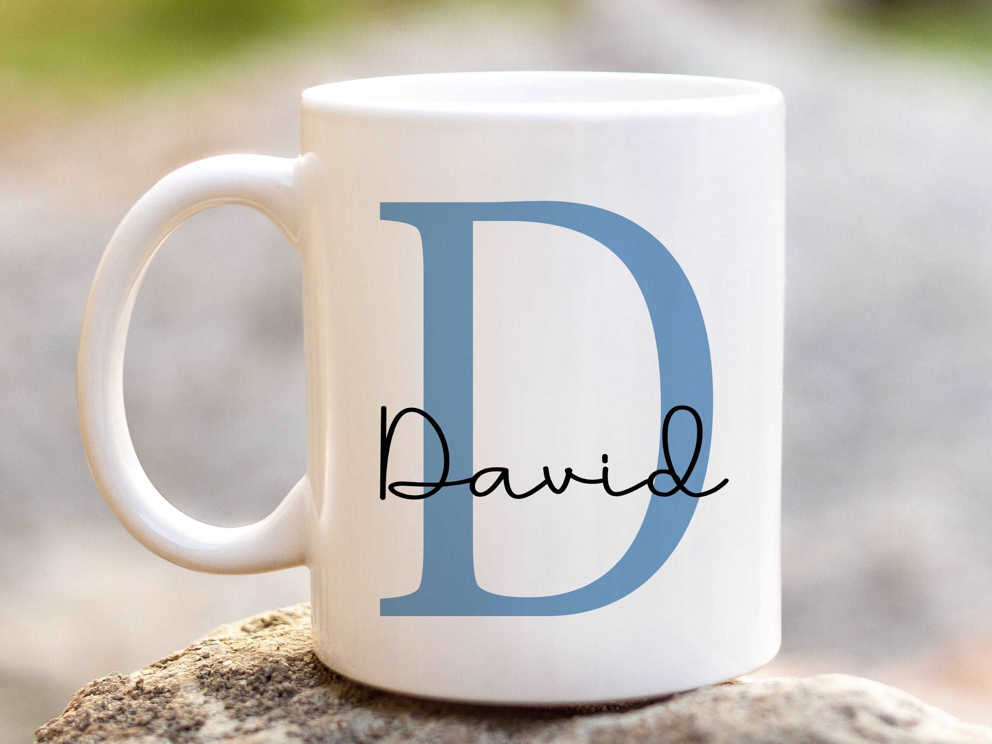 white mug sitting on a stone with a large blue letter D and the name David printed over in a script font